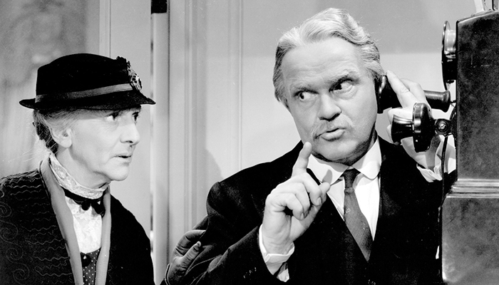 An older woman and man stand together, the man is speaking into a antique wall telephone.