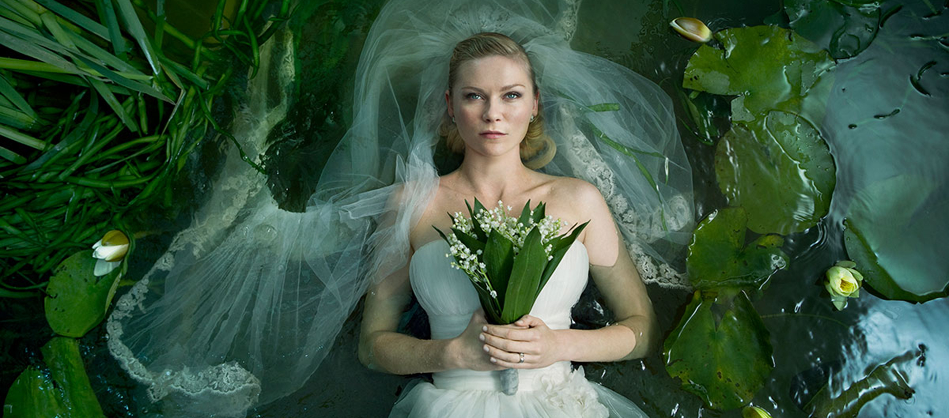 A woman in a wedding dress and veil lays in a pond surrounded by lily pads and reeds holding a bouquet, 