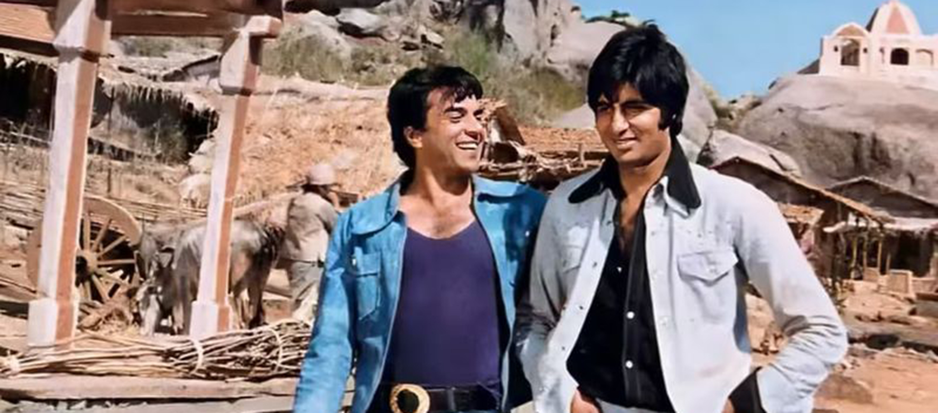 Two men stand together in a desert with stables and rocks in the background.