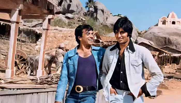 Two men stand together in a desert with stables and rocks in the background.