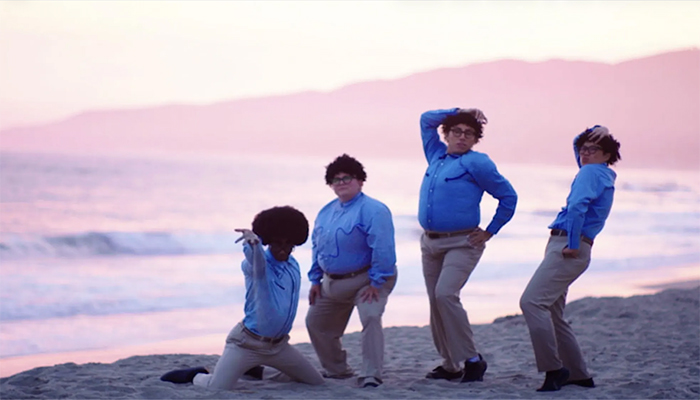 Four individuals pose on a beach wearing khaki pants, blue western shirts, glasses, and short curly-haired wigs.