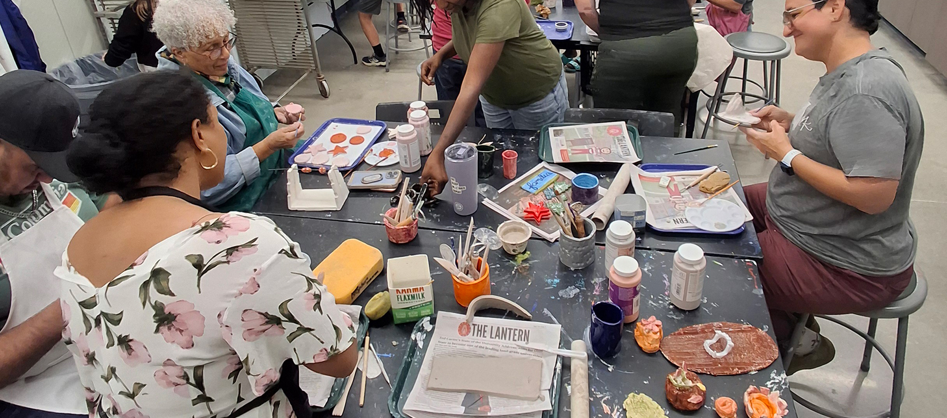 Five people sit and stand around a table with in-progress artwork made from clay slabs and art supplies. 