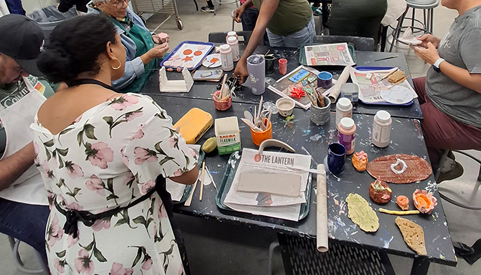 Five people sit and stand around a table with in-progress artwork made from clay slabs and art supplies. 