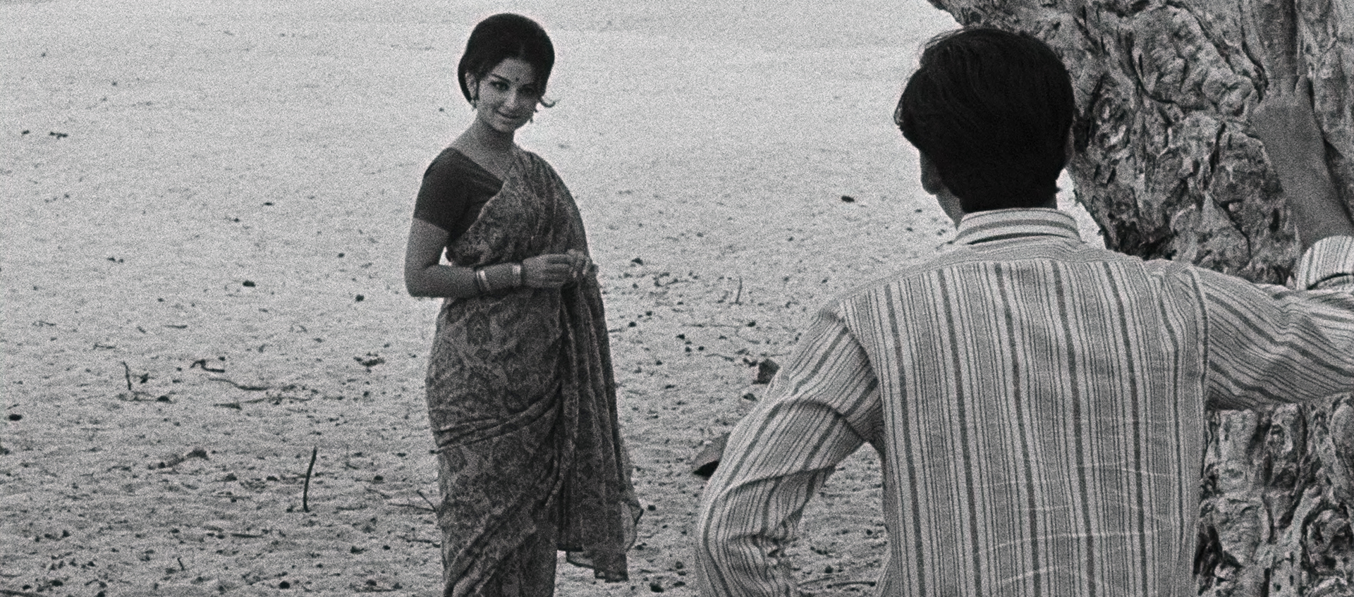 A woman wearing a sari stands in the sand, a man is facing her with his hand against a tree.