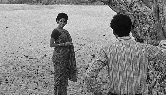A woman wearing a sari stands in the sand, a man is facing her with his hand against a tree.