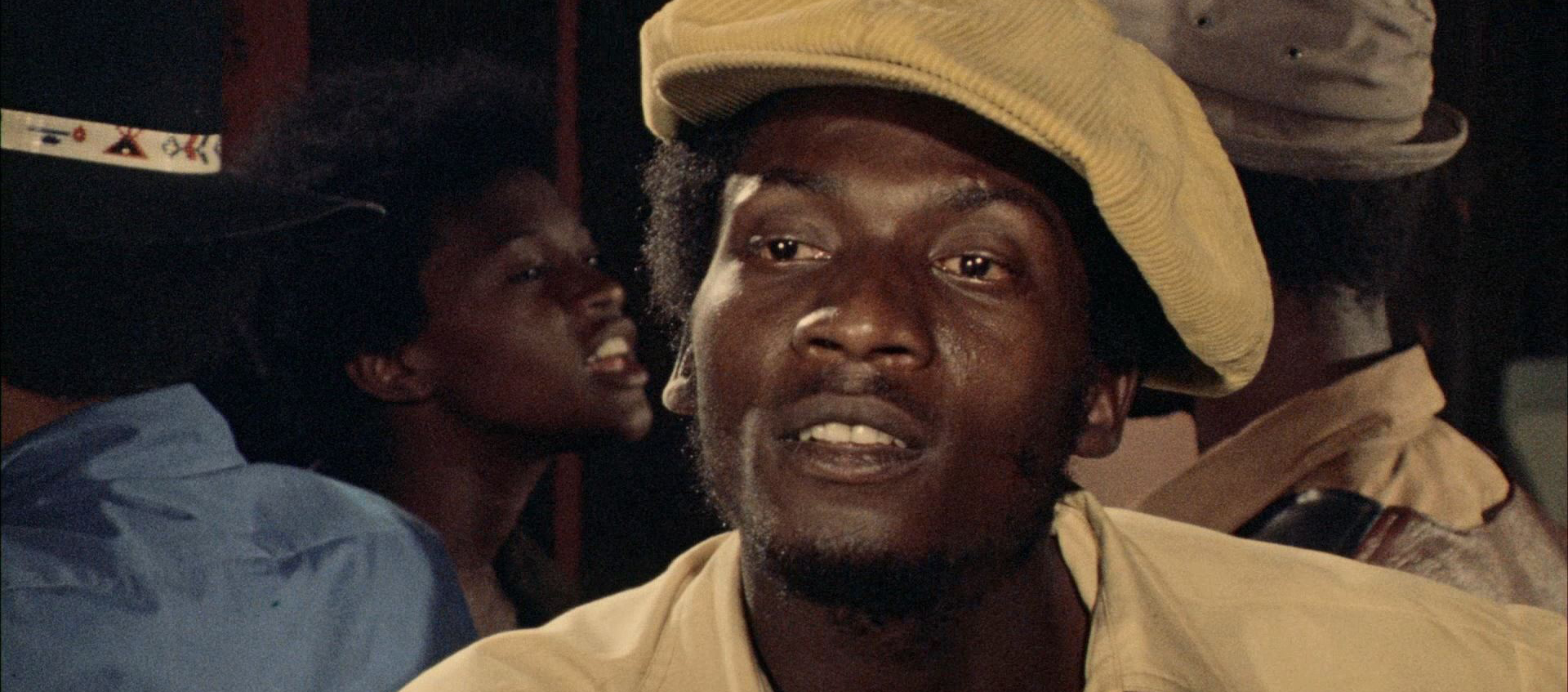 A man wearing a light yellow cap and shirt looks ahead, with other people visible in the background in a dimly lit setting.