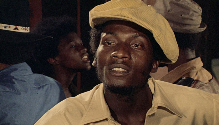 A man wearing a light yellow cap and shirt looks ahead, with other people visible in the background in a dimly lit setting.