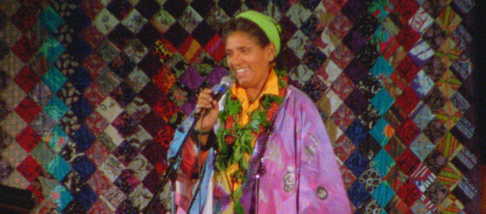A woman stands at a microphone wearing a colorful outfit and headband, a colorful quilt hangs behind her.