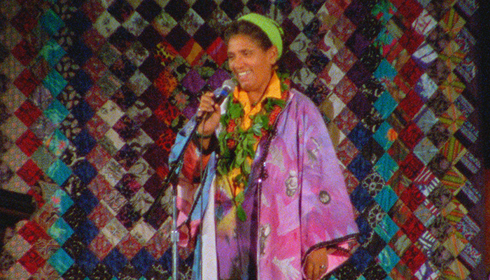A woman stands at a microphone wearing a colorful outfit and headband, a colorful quilt hangs behind her.