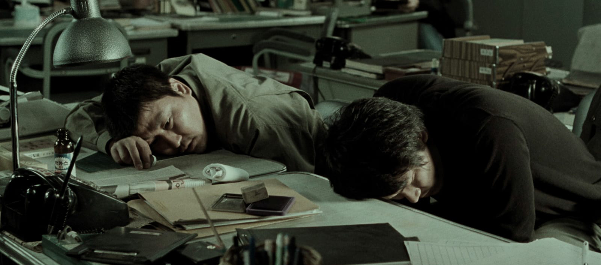 Two men lay their heads on a desk covered with papers in a dimly lit office.