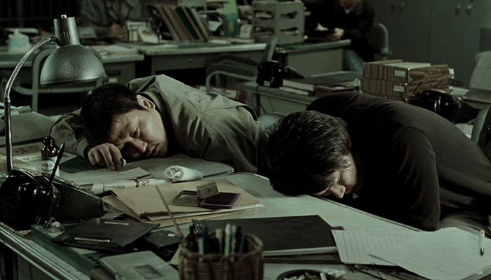 Two men lay their heads on a desk covered with papers in a dimly lit office.