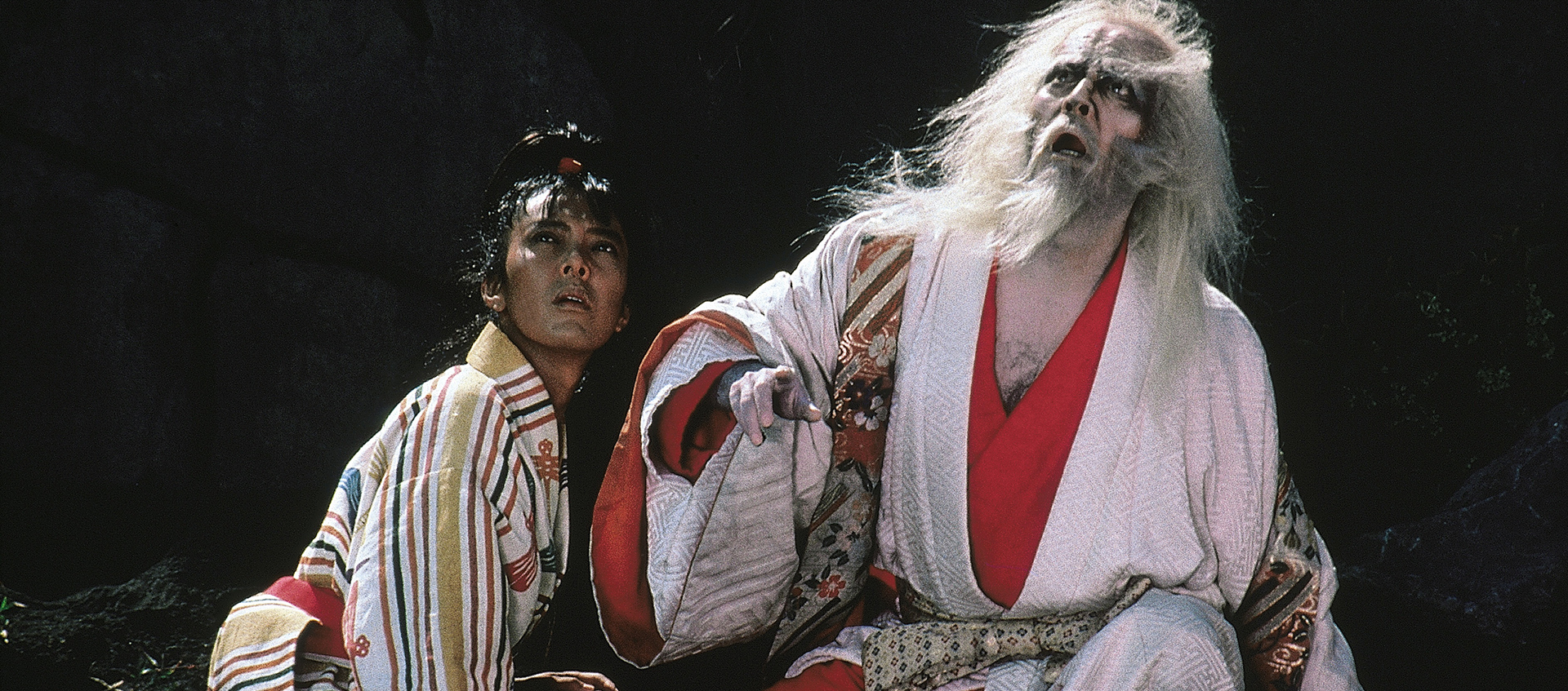 Two individuals in kimonos kneel on the ground together looking up at something.