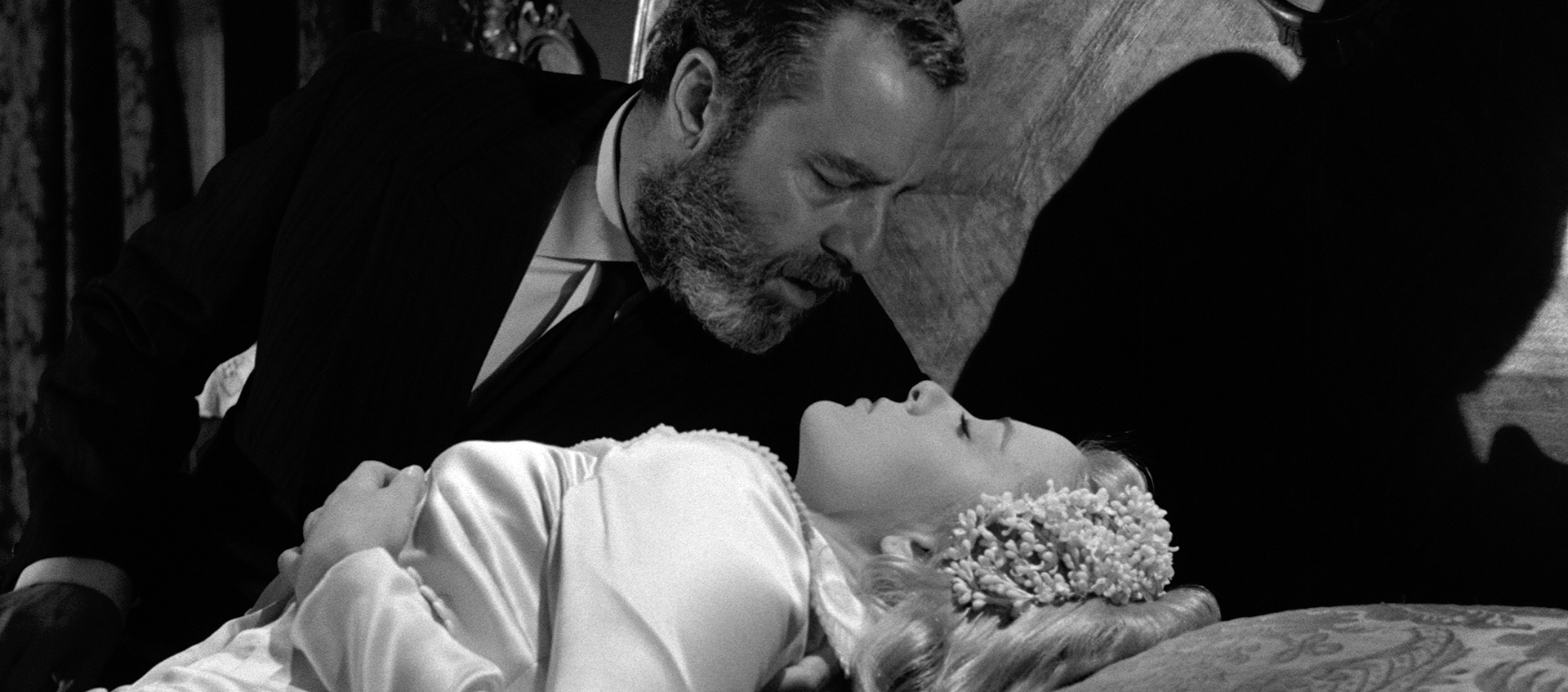 A bearded man leans over a woman in a wedding dress who is lying down on a bed, in a black and white scene.