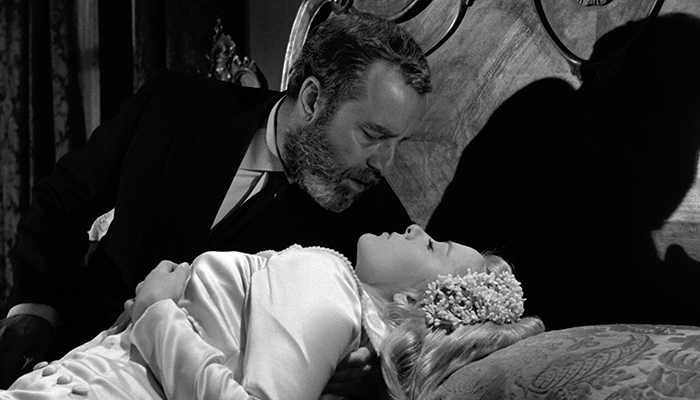 A bearded man leans over a woman in a wedding dress who is lying down on a bed, in a black and white scene.