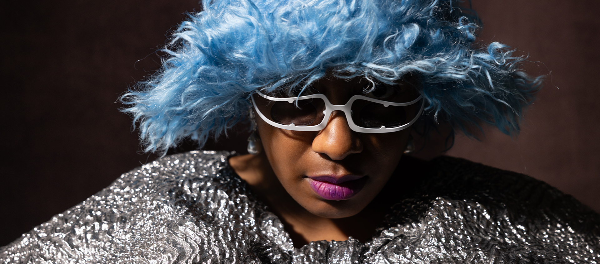 A woman wears a fuzzy blue hat, white elongated sunglasses and a textured silver cape
