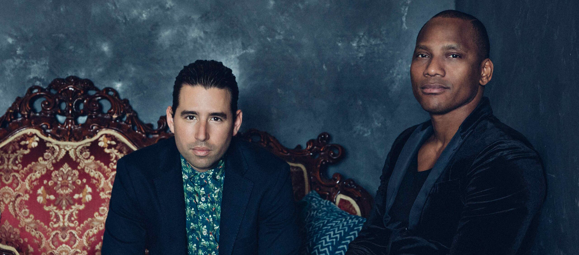 two men in suits sit side by side on an ornate couch