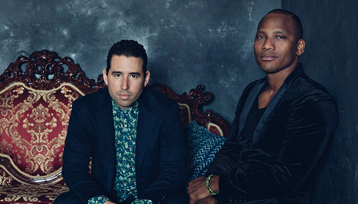 two men in suits sit side by side on an ornate couch