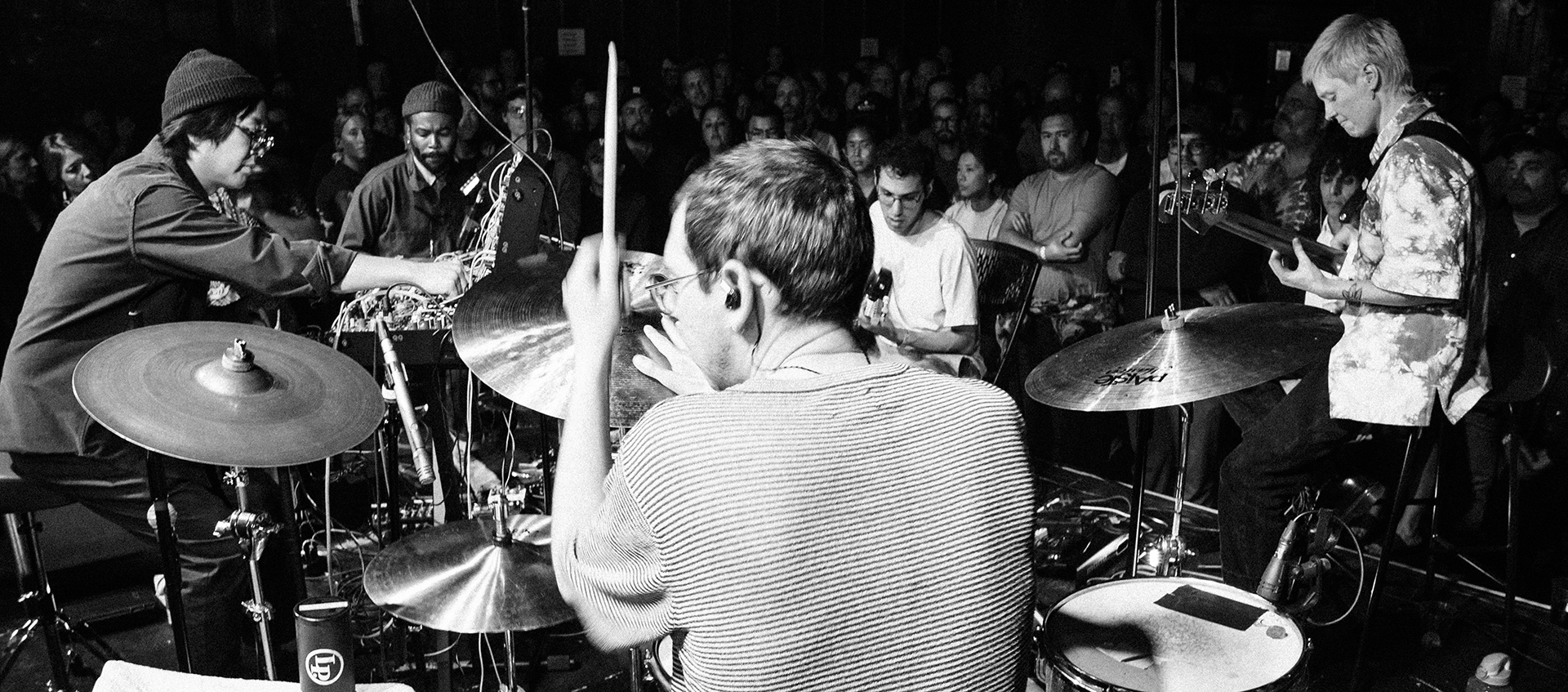 view of man playing a drumset from behind with other musicians and crowd in front
