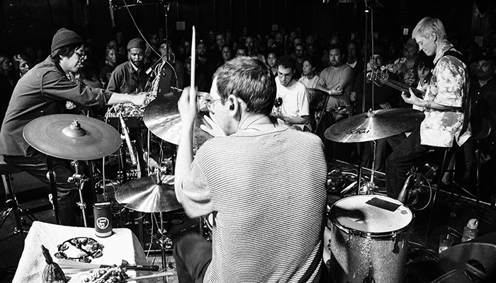 view of man playing a drumset from behind with other musicians and crowd in front