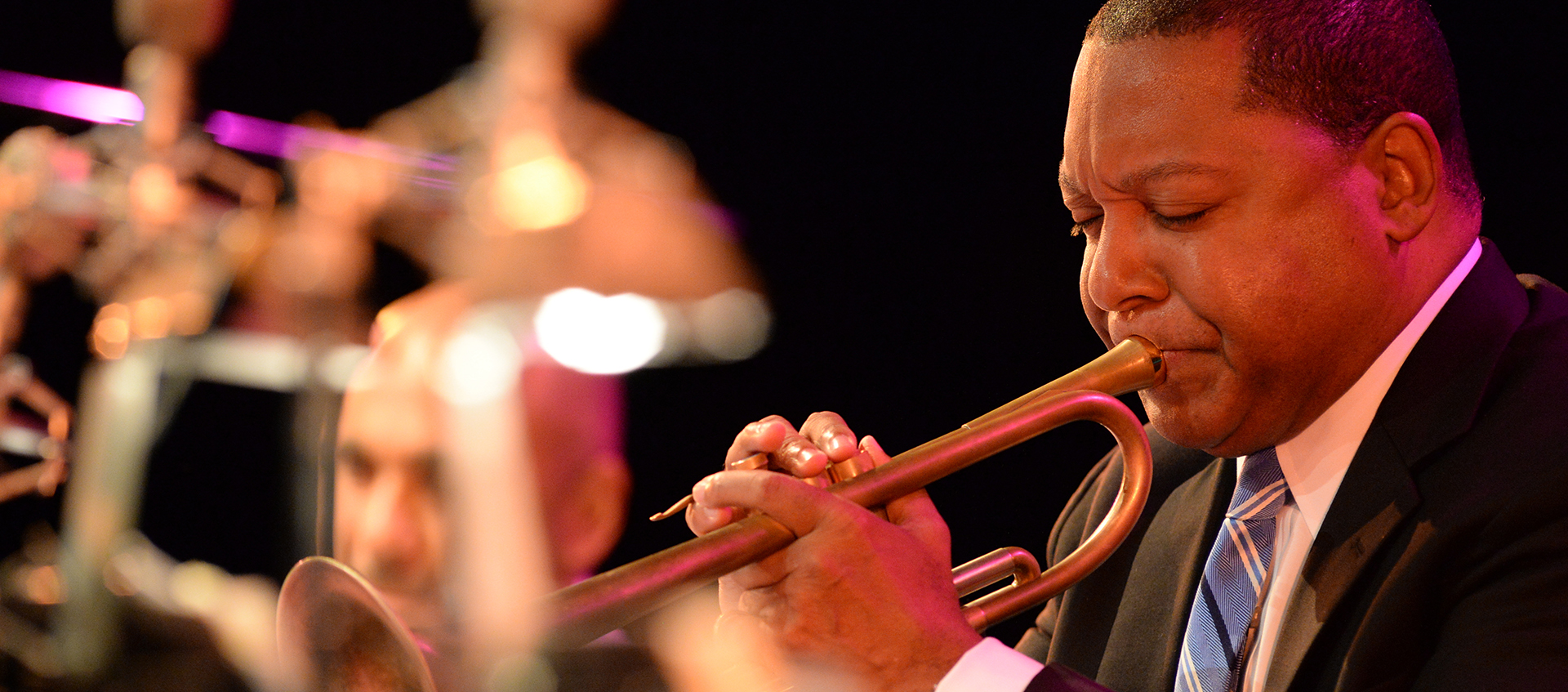 A profile view of Wynton Marsalis playing the trumpet. Members of his band are blurred in the background.