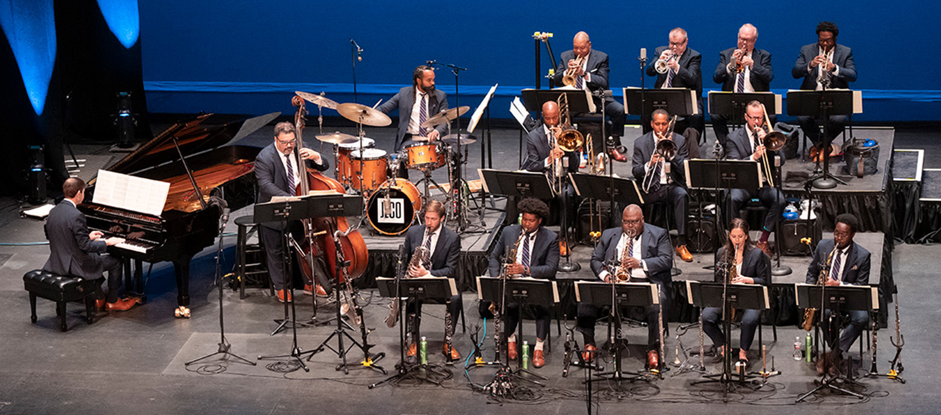 A jazz orchestra sits holding instruments on a stage.