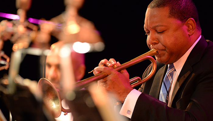 A profile view of Wynton Marsalis playing the trumpet. Members of his band are blurred in the background.