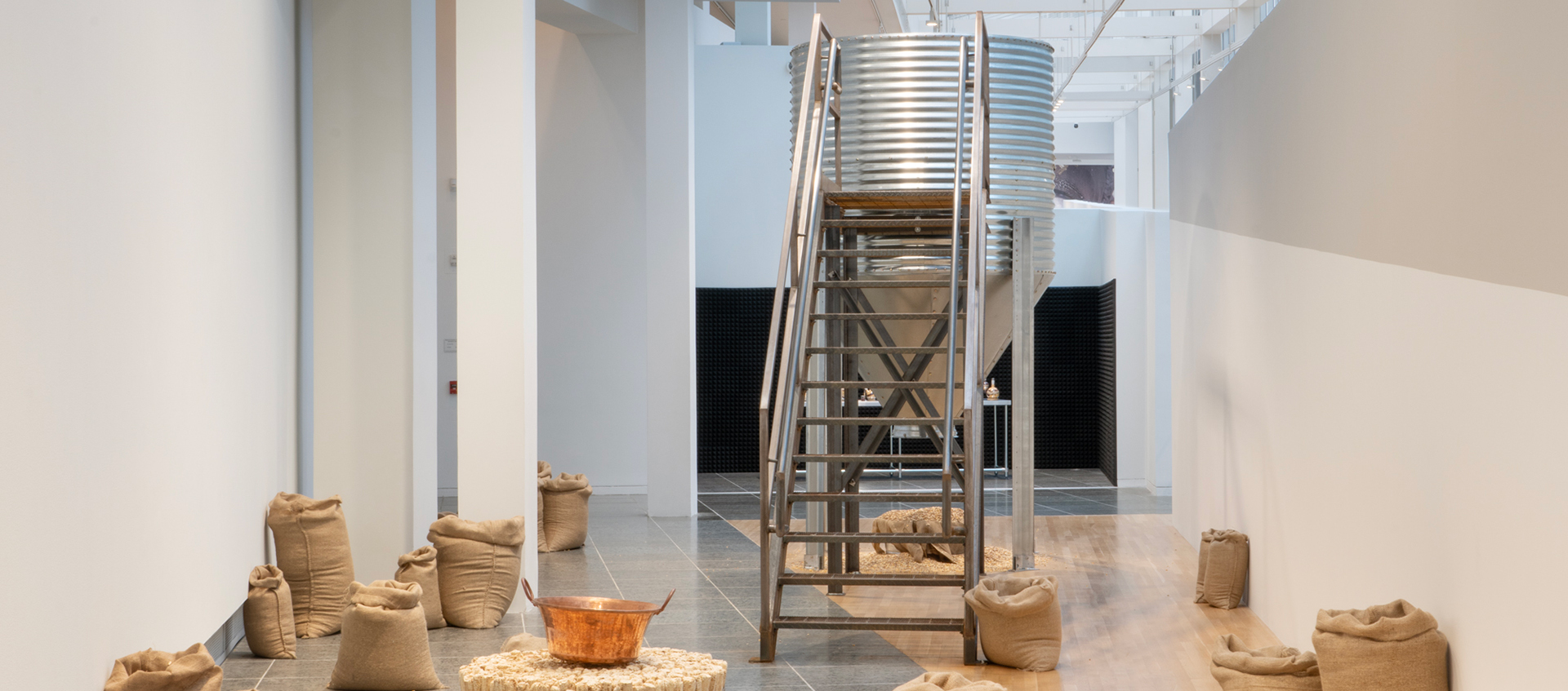 Burlap sacks and a table made from corn cobs flank a metal silo that has a staircase attached to it and a pile of seeds below it. 