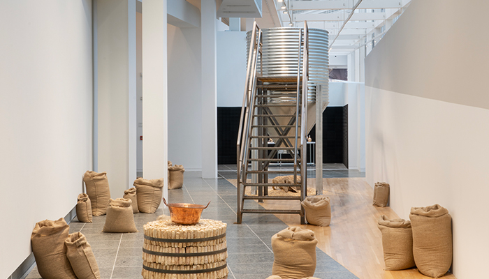 Burlap sacks and a table made from corn cobs flank a metal silo that has a staircase attached to it and a pile of seeds below it. 