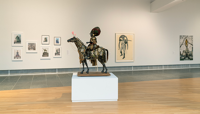 A black equestrian statue of a man in gold-ornamented armor and a large headpiece. The horse is adorned with flowers, gold chains, and medallions.
