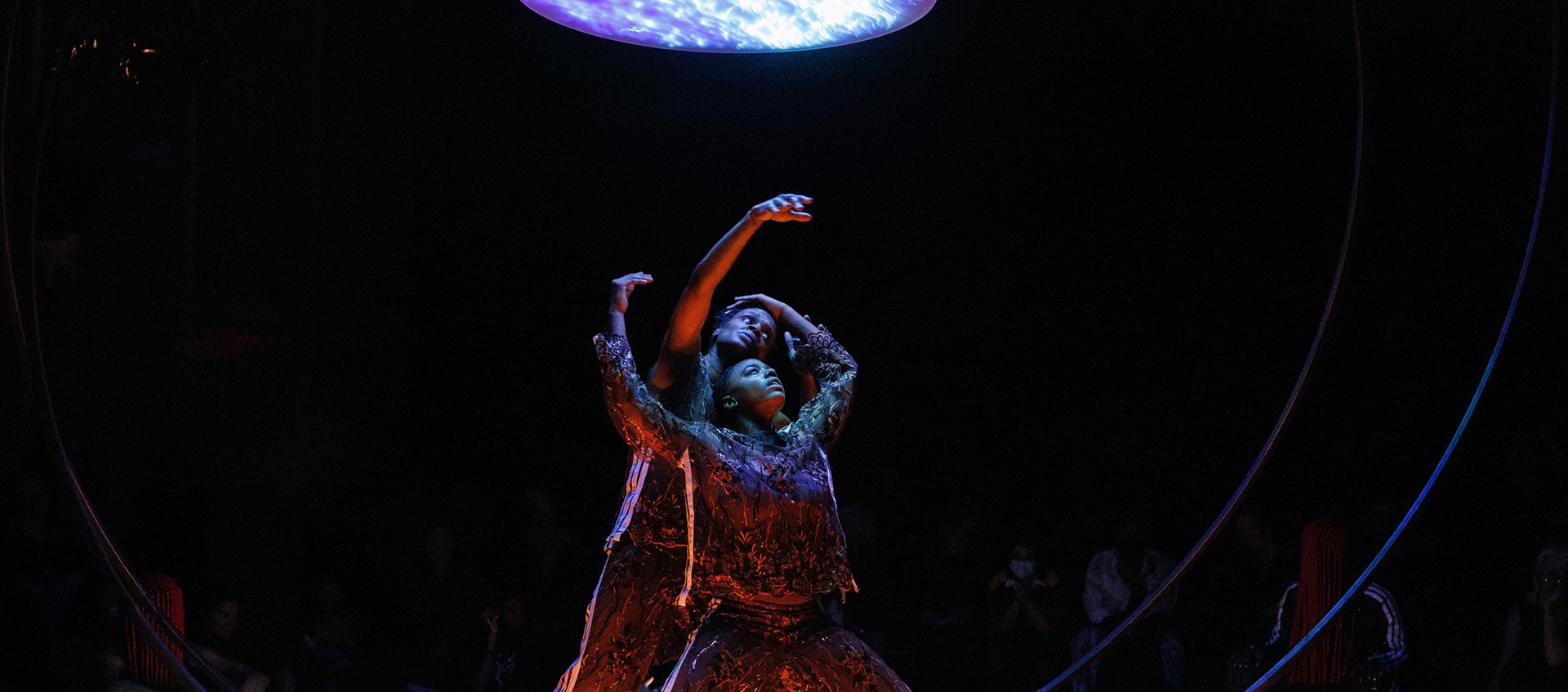 Two female dancers stand between two vertical metal rings on a dark stage illuminated from above by a blue disk.