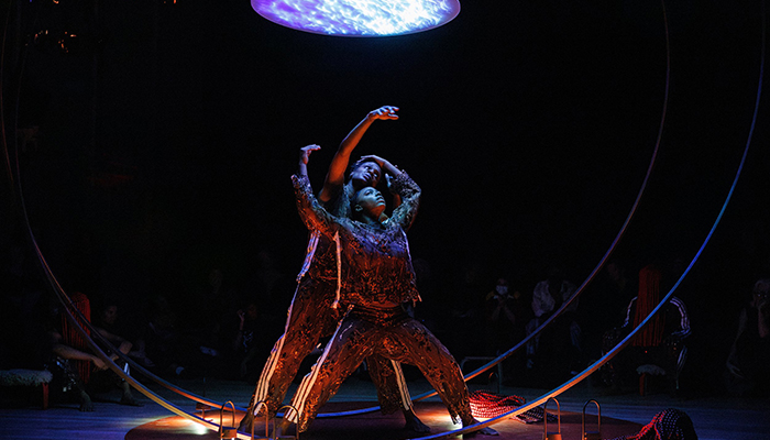 Two female dancers stand between two vertical metal rings on a dark stage illuminated from above by a blue disk.