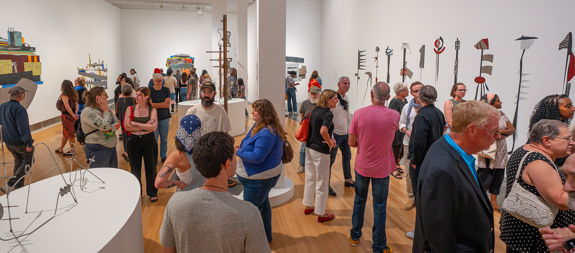 A large group of people gathered in a gallery look at the artwork on the walls and pedestals and converse with one another. 
