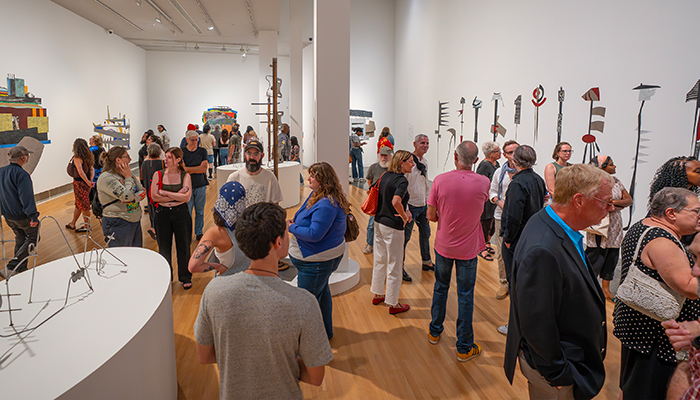 A large group of people gathered in a gallery look at the artwork on the walls and pedestals and converse with one another. 