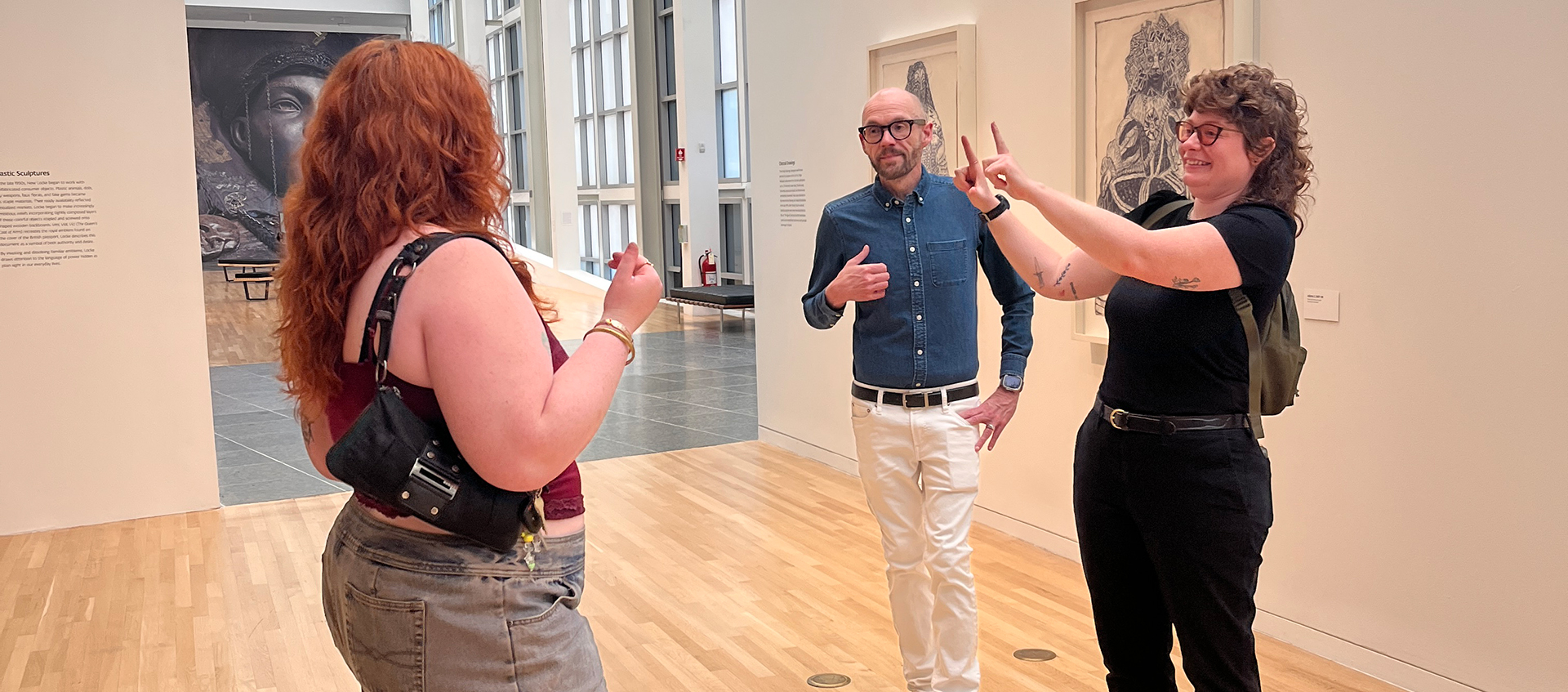 Three people stand in a gallery and make gestures with their hands while looking at one another. 