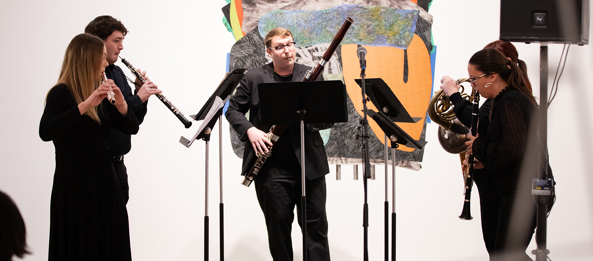 Four musicians play wind and reed instruments and look toward their music stands while standing in a gallery. 