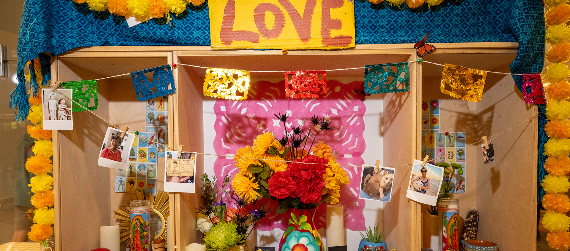 Altar with paper and flower garlands, photos, candles, calaveras, vases, flowers, postcards, and a sign reading "Love."