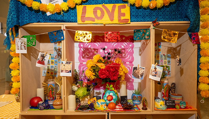 Altar with paper and flower garlands, photos, candles, calaveras, vases, flowers, postcards, and a sign reading "Love."