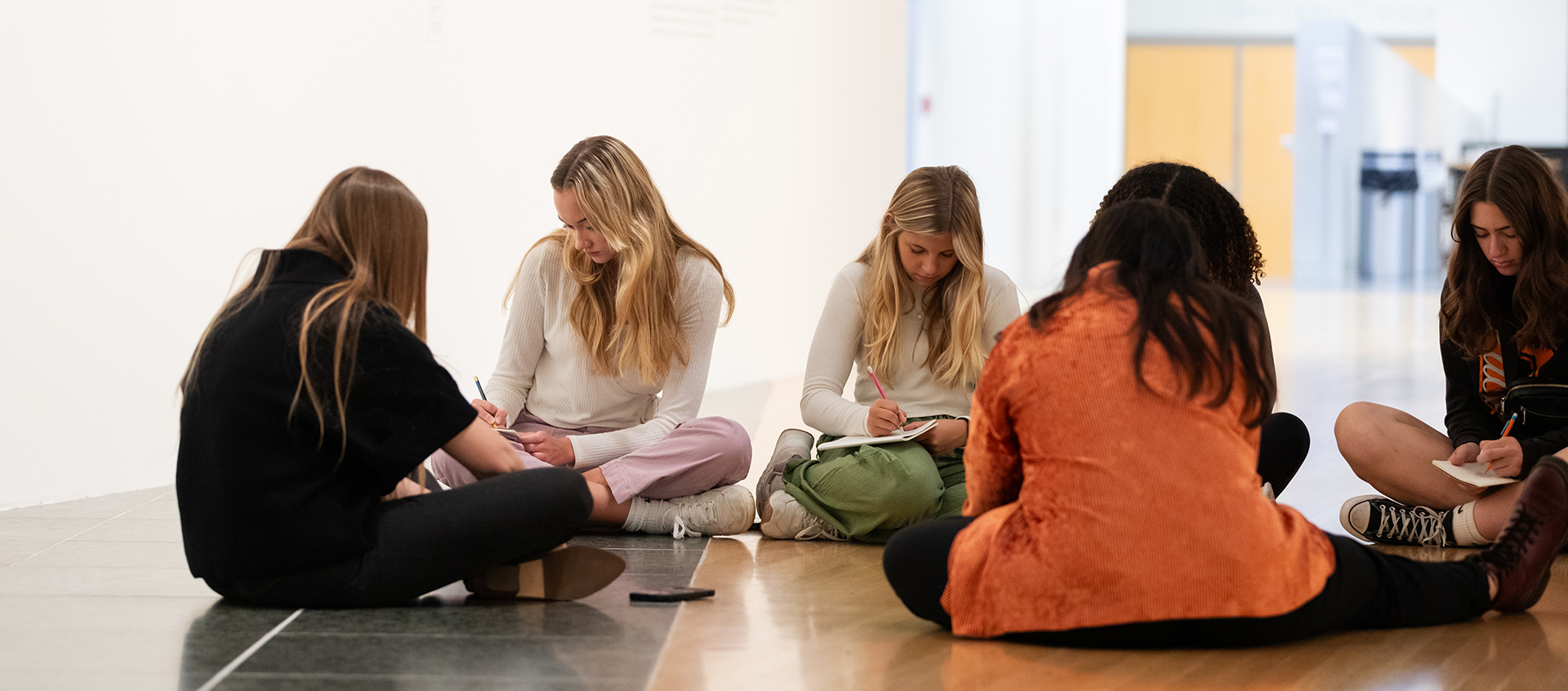 A group of 5 people sit in a circle on a gallery floor. Many hold notebooks in their hands.