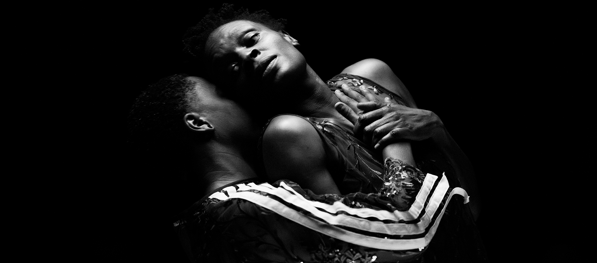 Two people embrace closely in dramatic lighting against a dark background, one with eyes closed and the other gazing upward.