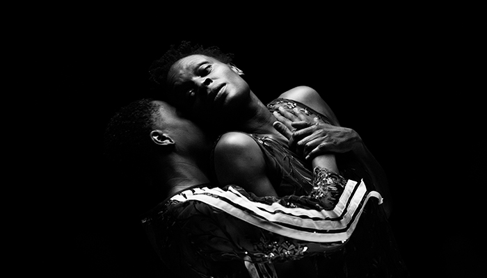 Two people embrace closely in dramatic lighting against a dark background, one with eyes closed and the other gazing upward.
