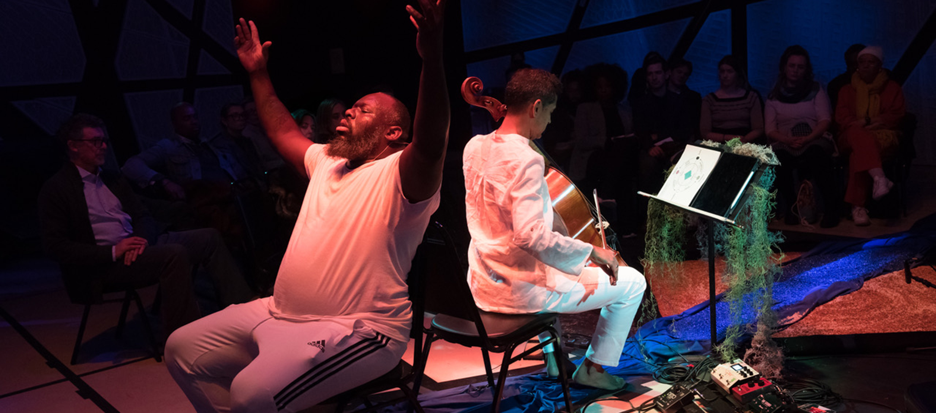 Two men sit back to back, one has his hands outstretched the other is playing a cello.