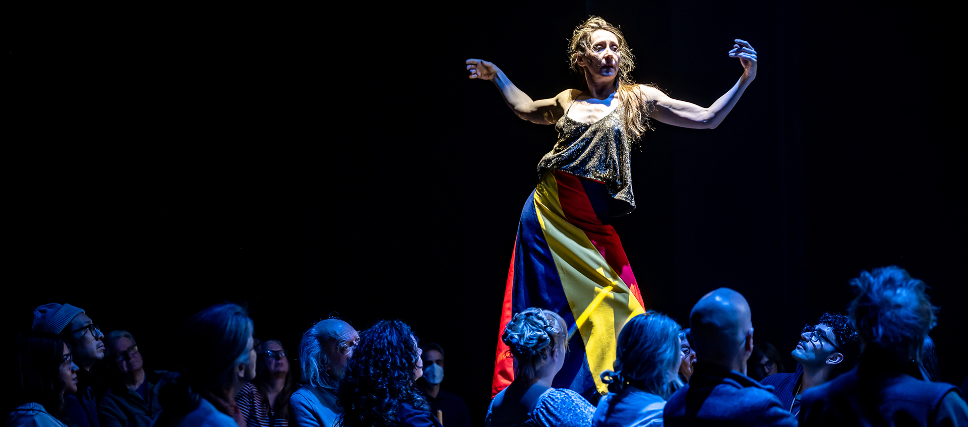 A woman stands with her arms outstretched on a table with audience members gathered around.
