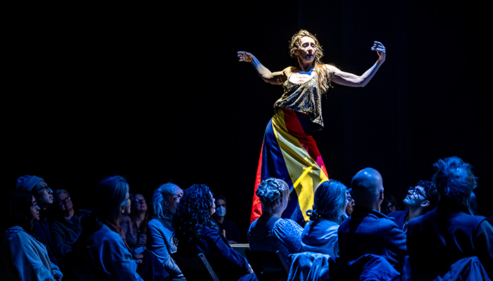 A woman stands with her arms outstretched on a table with audience members gathered around.