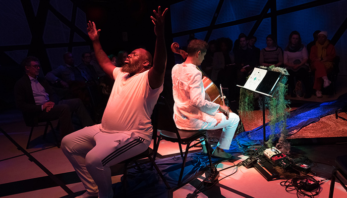 Two men sit back to back, one has his hands outstretched the other is playing a cello.