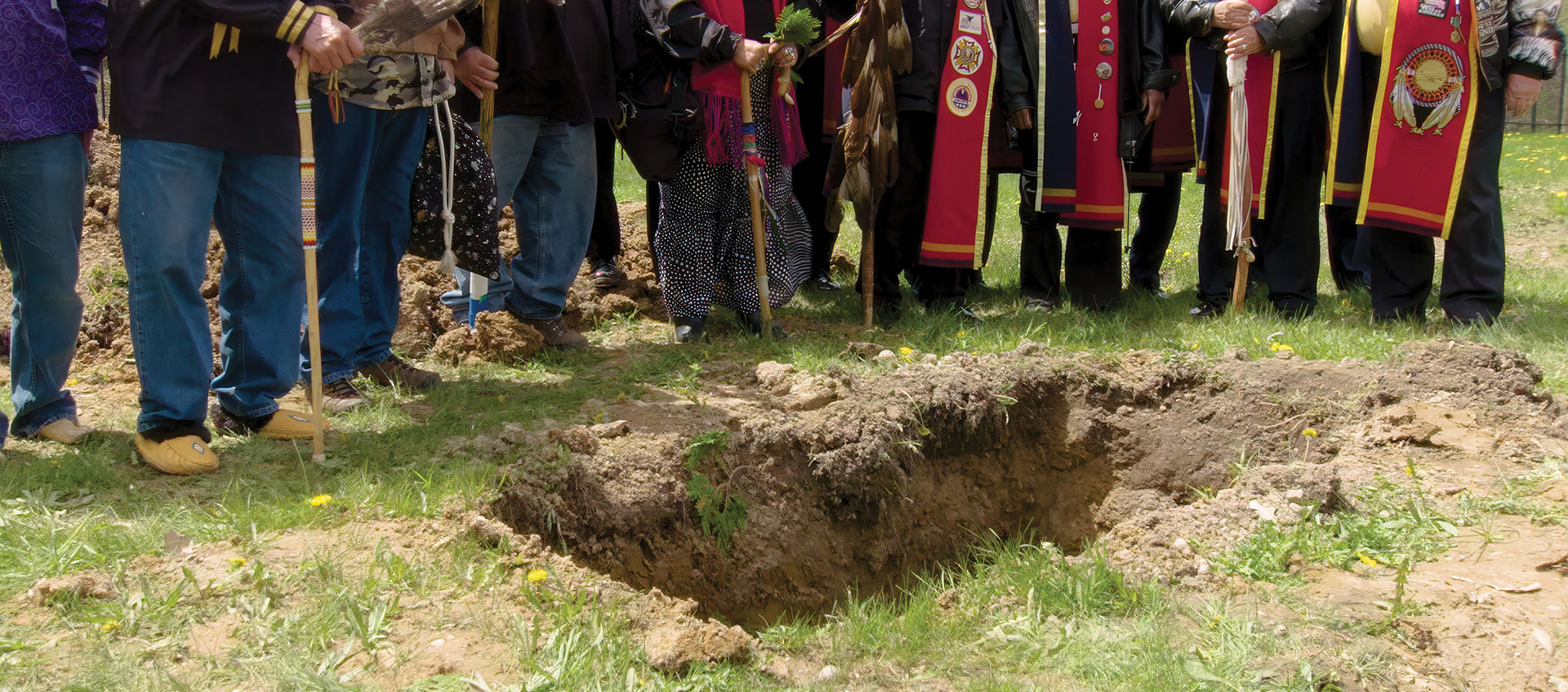 A group of people wearing regalia stand around an empty grave.