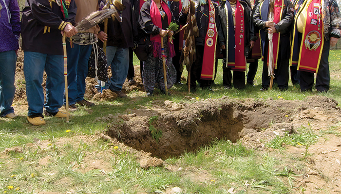 A group of people wearing regalia stand around an empty grave.