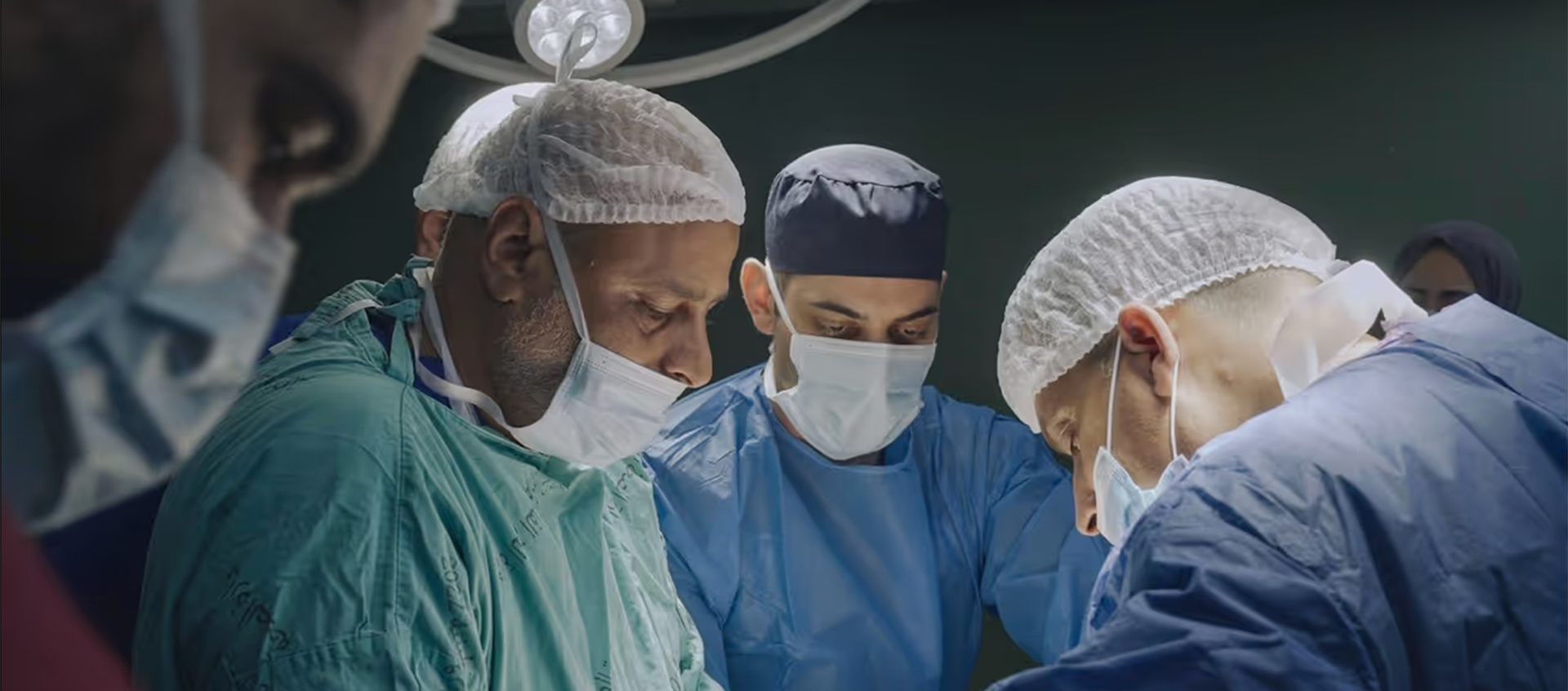 Three doctors wear scrubs and masks in an operating room.