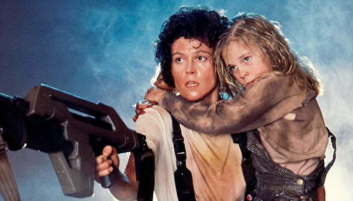 A woman with a dirty white t shirt holds a child and a large gun.