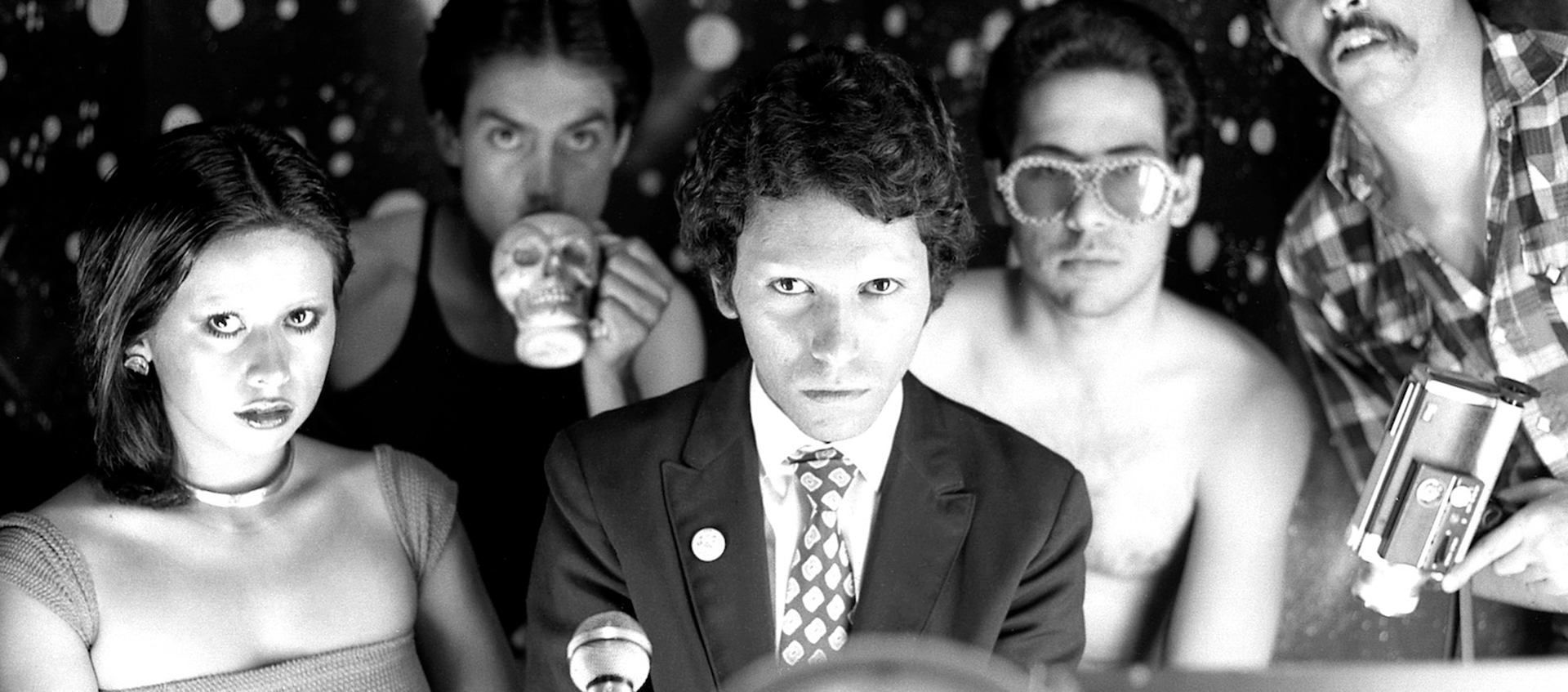 Five people stand together, one holds a camera, one has large sunglasses, two do not have eyebrows, and one is holding a skull.