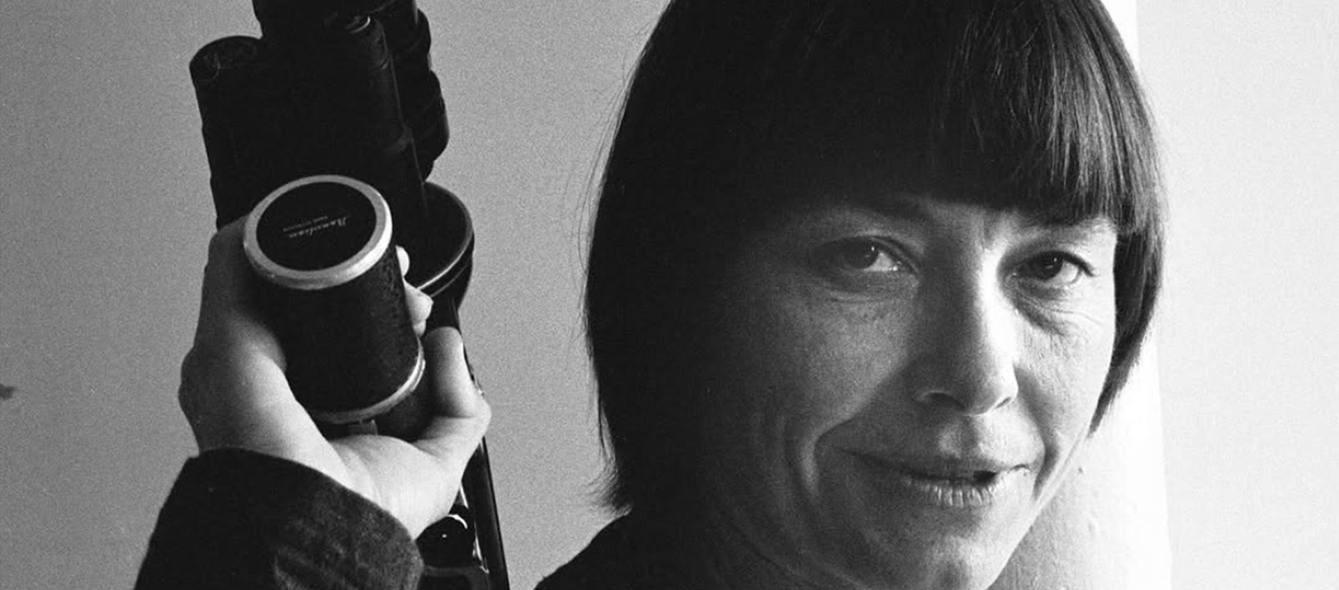 A woman with short hair and bangs holds a camera beside her head.